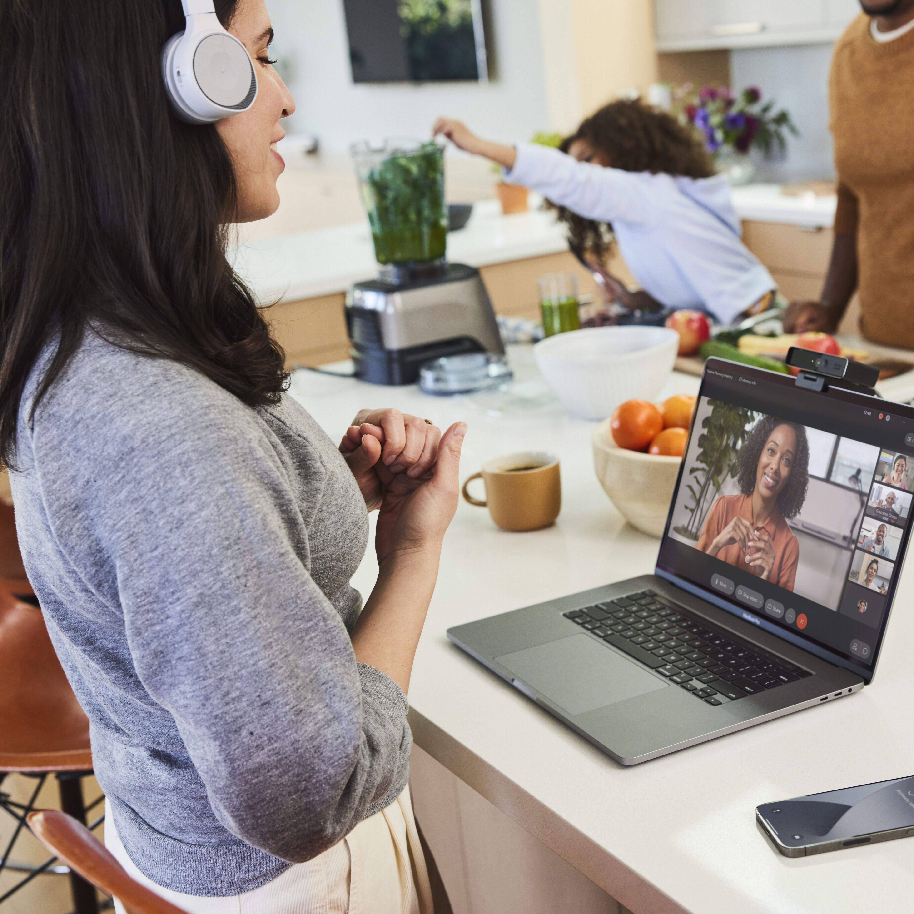 Person in a headset video conferences from their kitchen while their partner and child make a smoothie in the background.